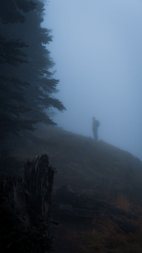 Silhouette of a Man with a Backpack Standing on a Hill Slope in Fog