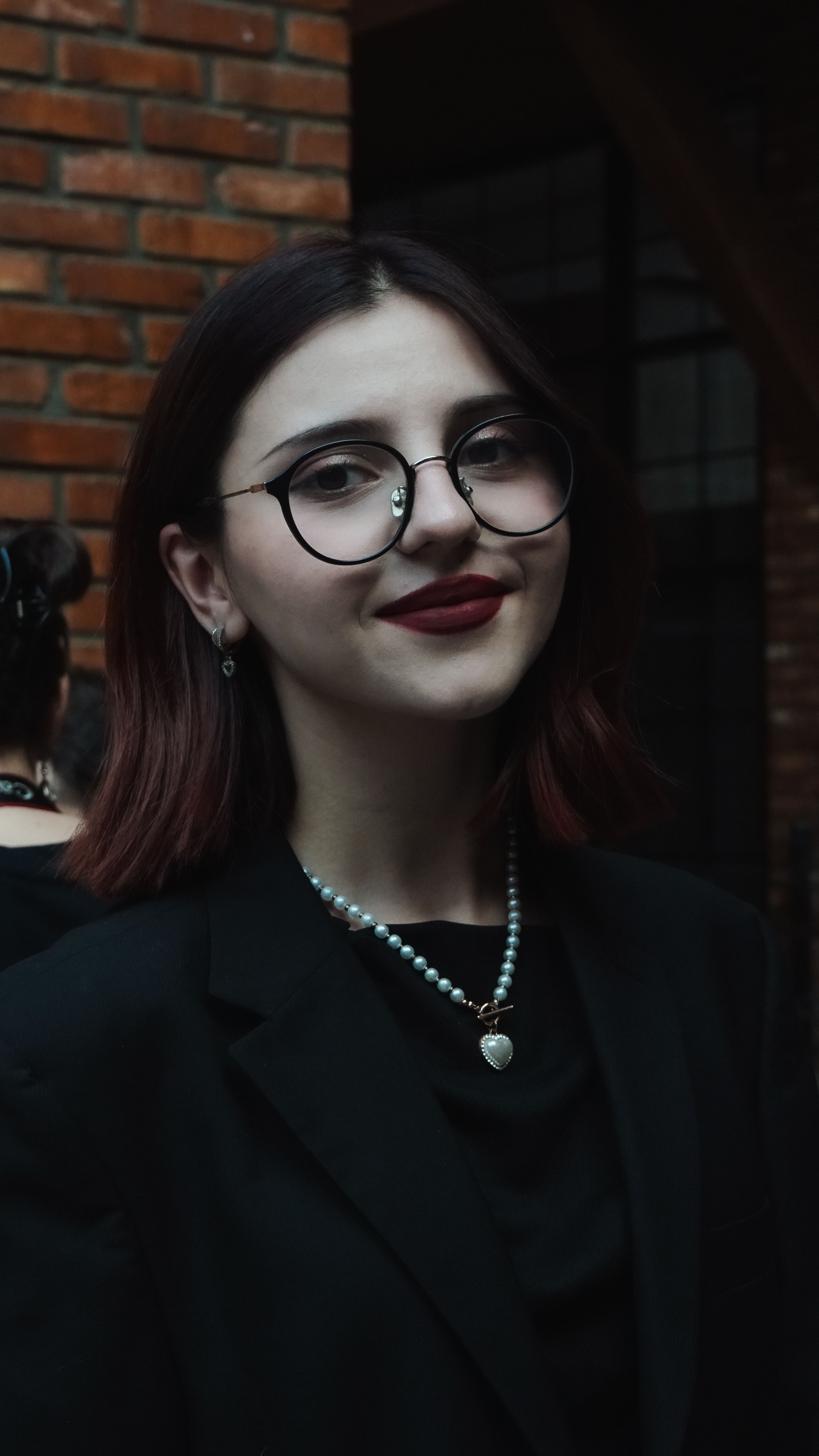 Portrait of a fashionable young woman in glasses, wearing a black blazer and pearl necklace.