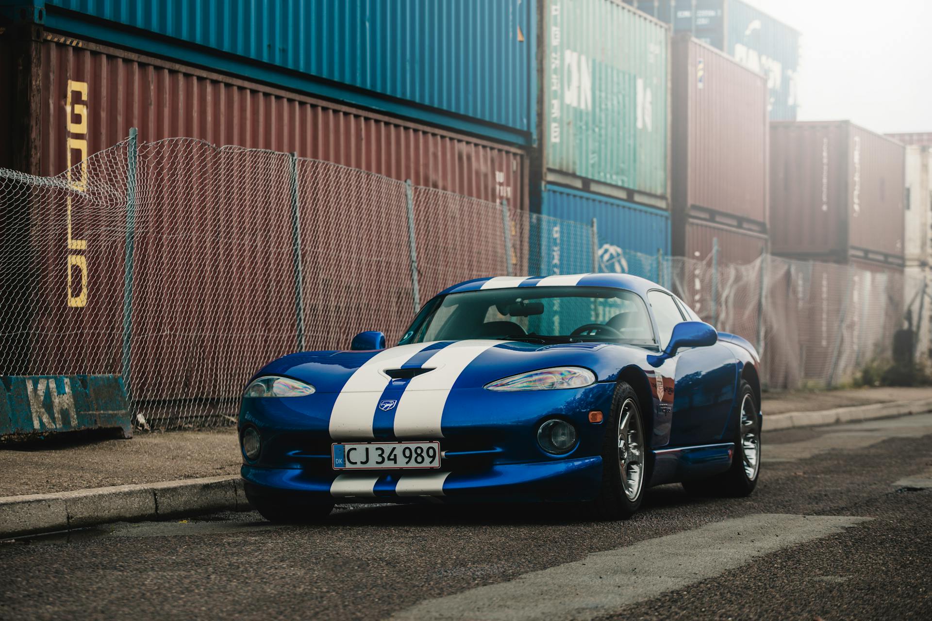 A sleek blue sports car parked on city streets surrounded by shipping containers in København.