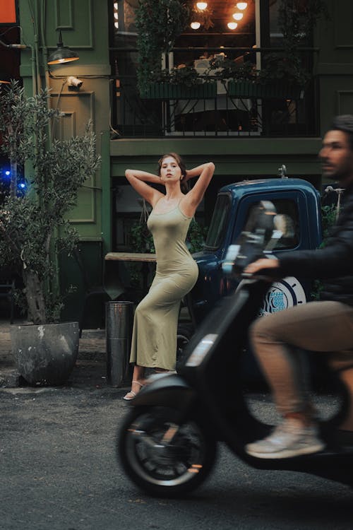 Model in Green Dress Standing on Street behind Man Riding Motor Scooter