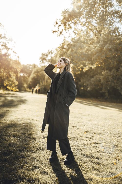 Free Woman in elegant coat posing in a sunlit park during fall, exuding style and grace. Stock Photo