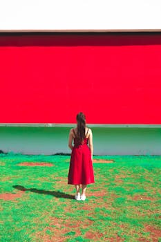 一名身穿红色连衣裙的女子站在绿地上，面对着一面鲜艳的红墙，形成了鲜明的对比