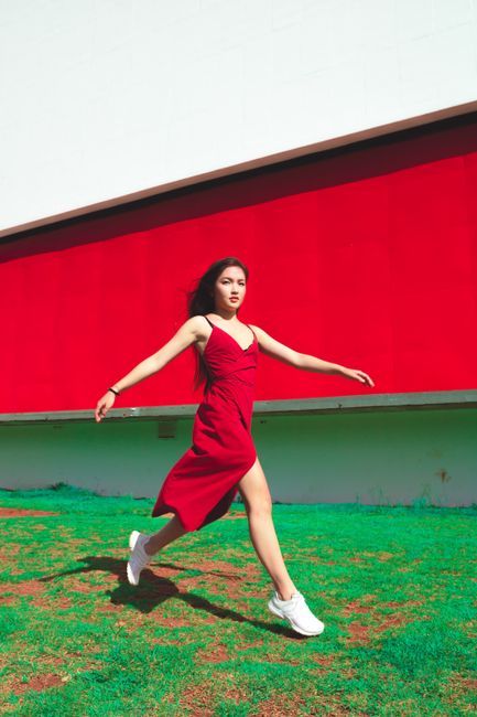 Energetic woman in red dress runs outdoors against bold red wall.