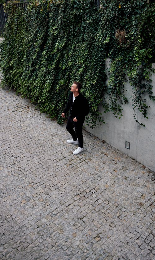 A Man Standing on a Pavement by the Wall Covered with a Climbing Plant 