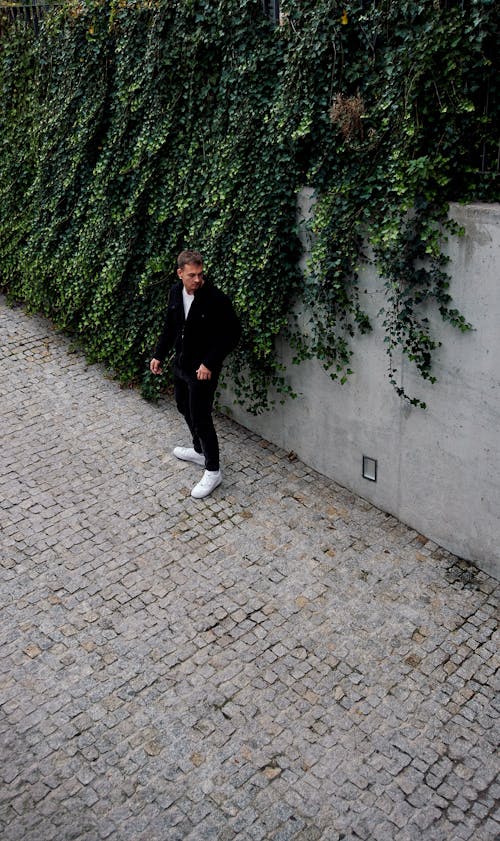 A Man Standing on a Pavement by the Wall Covered with a Climbing Plant 