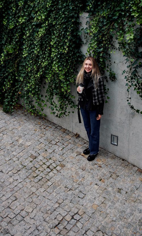 A Woman Standing on a Pavement by the Wall Covered with a Climbing Plant 