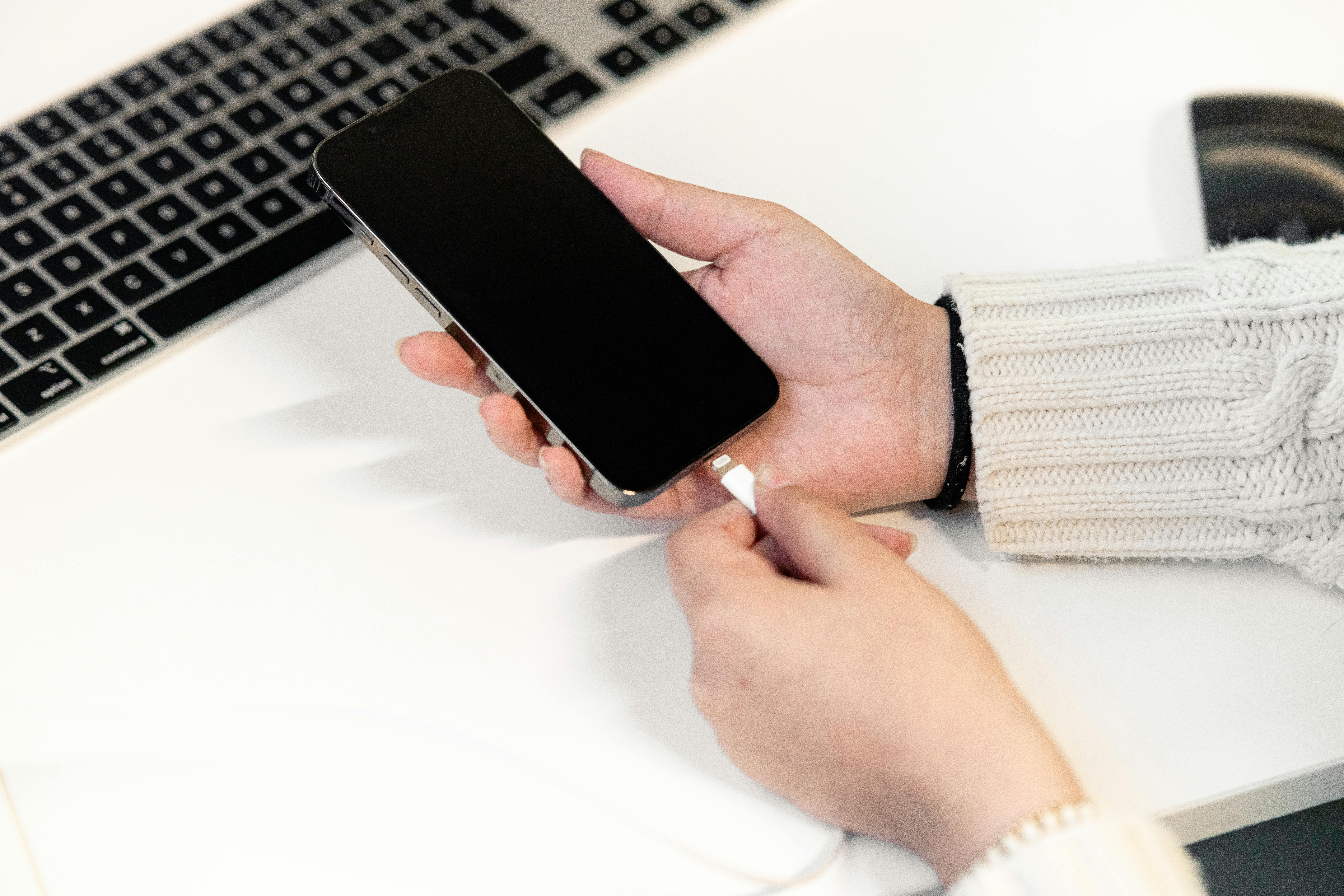 A woman plugs her smartphone into a charger at a desk, next to a laptop, illustrating modern technology usage.