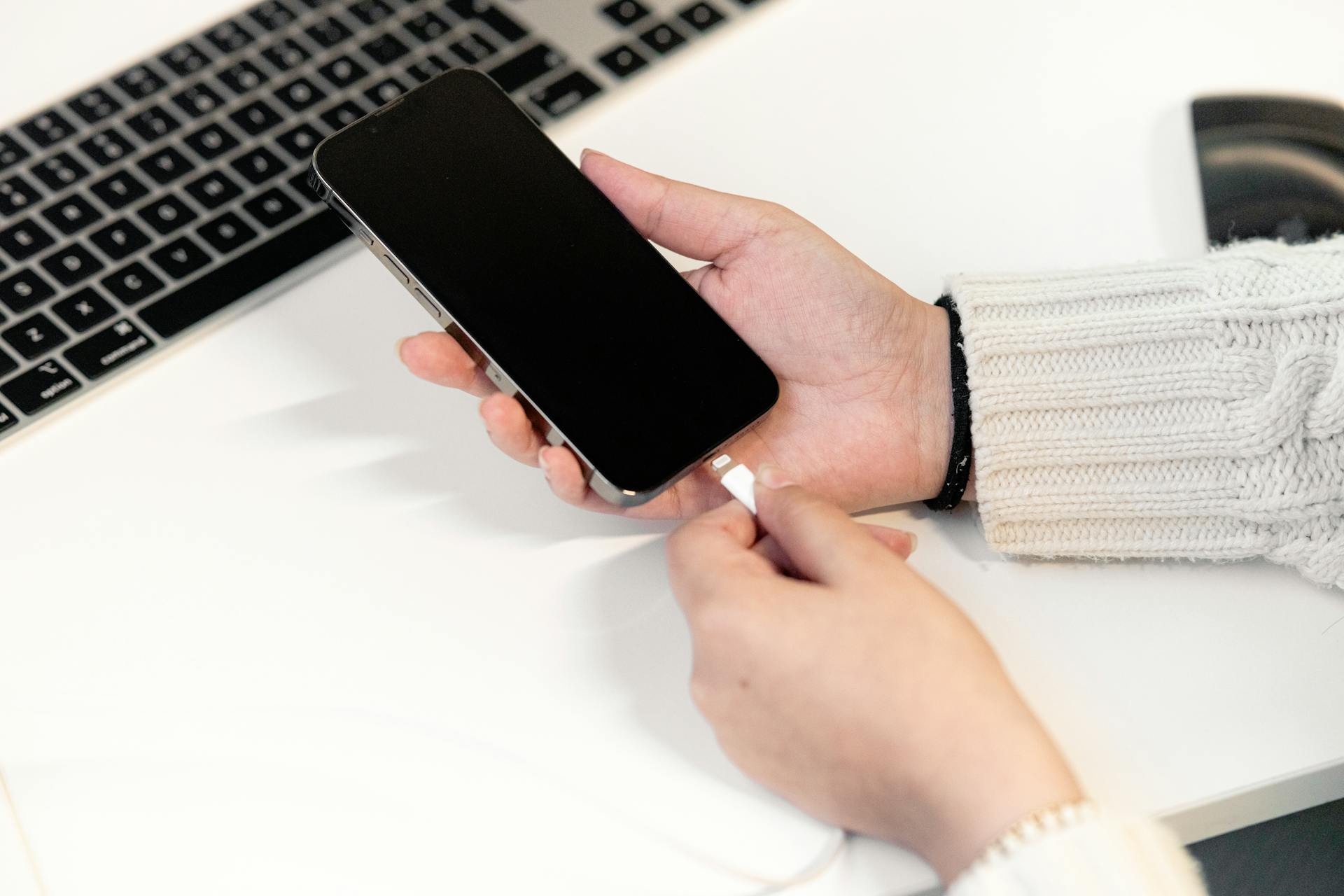 A woman plugs her smartphone into a charger at a desk, next to a laptop, illustrating modern technology usage.