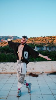 A carefree man in stylish fashion dances on a terrace with a scenic mountainous backdrop at sunset.