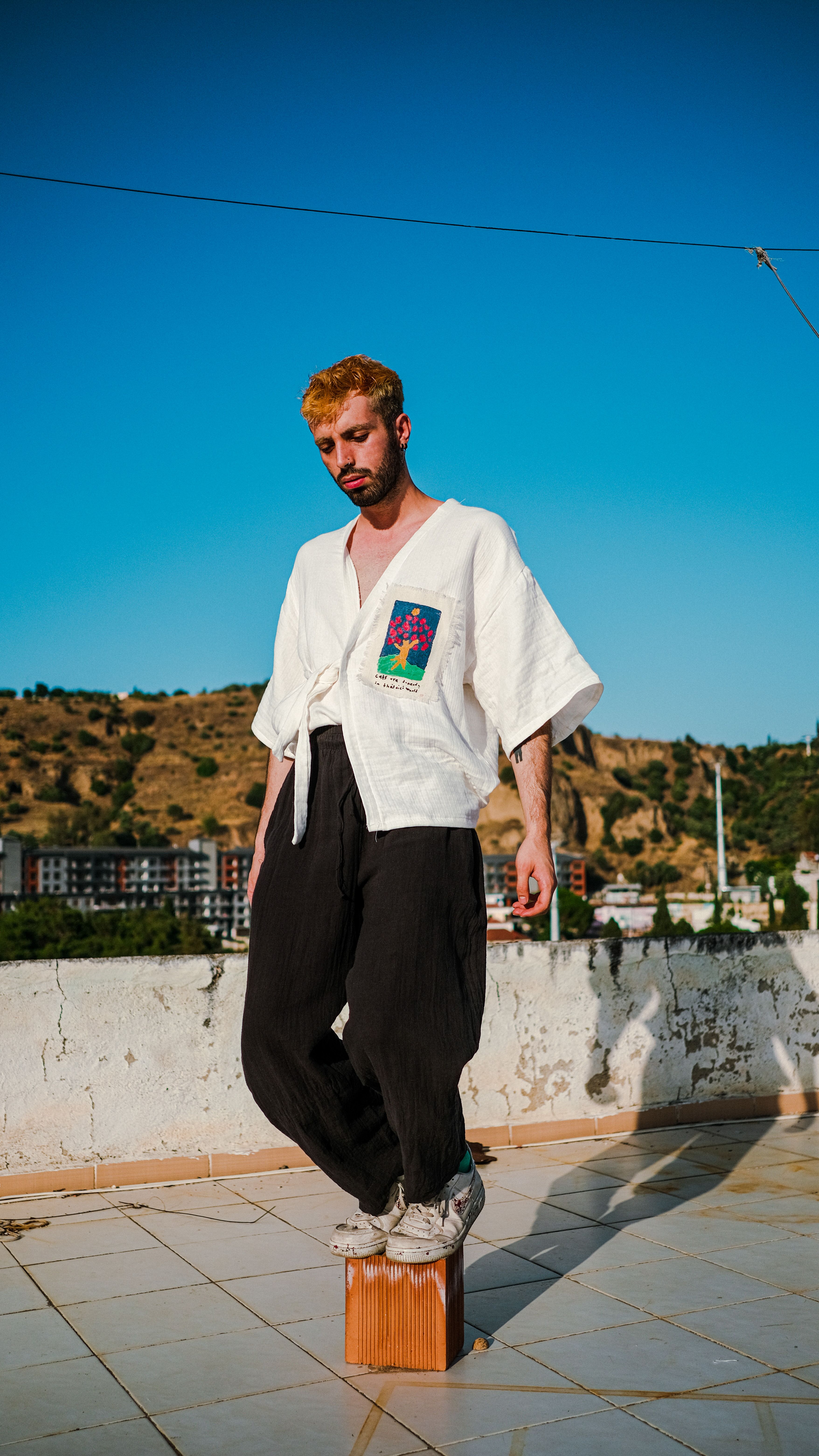 Stylish man in white shirt and black pants posing on a rooftop under clear blue sky.