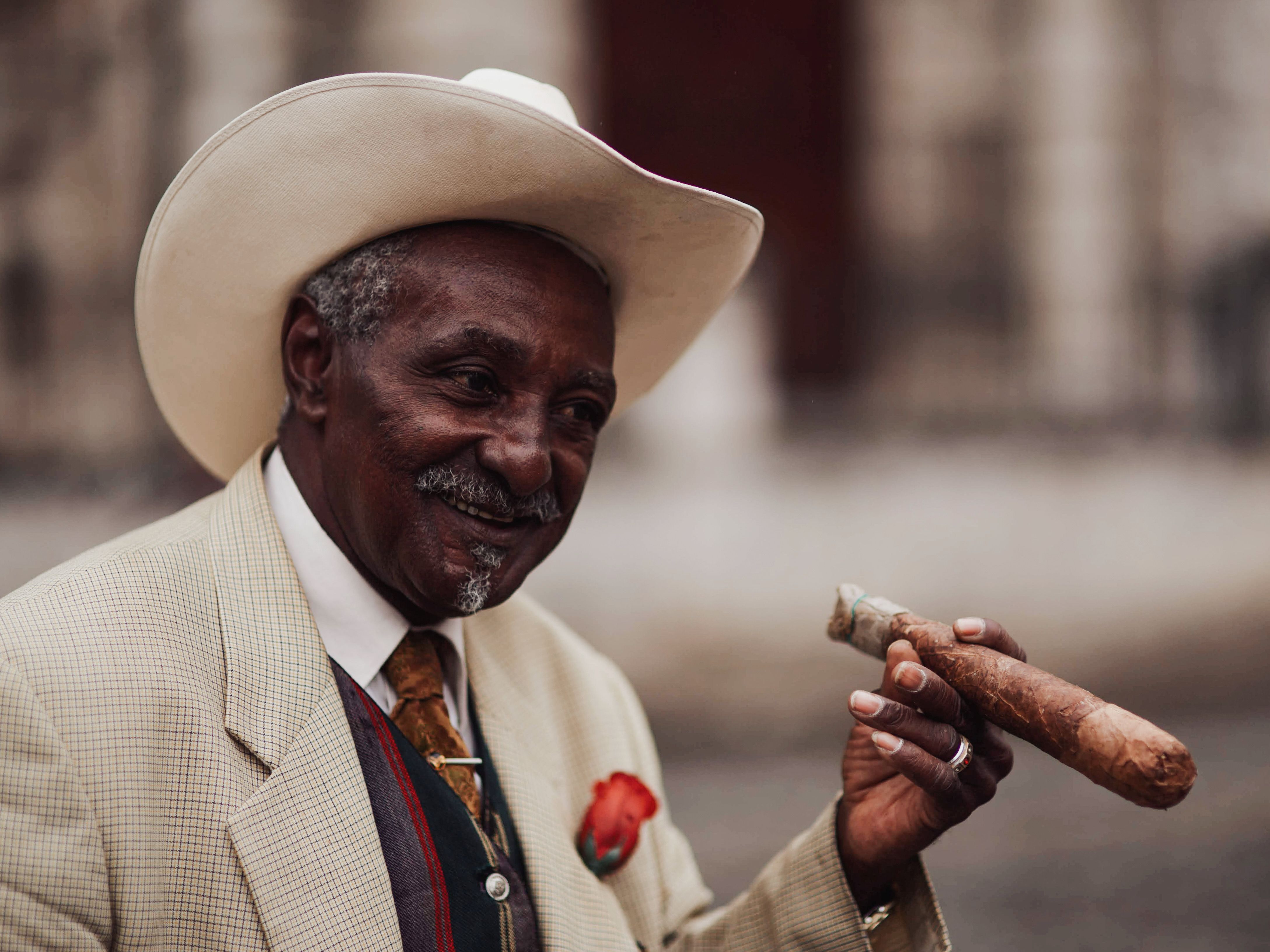 Portrait of a senior man in a cowboy hat and suit, smiling while holding a cigar outdoors.