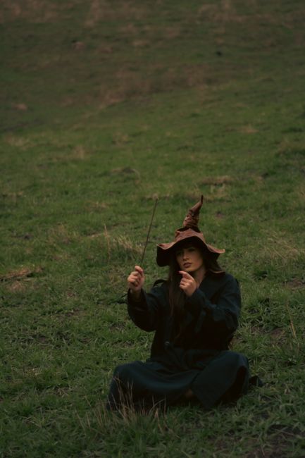 A woman dressed as a witch sits on a grassy hill, casting spells with a wand.