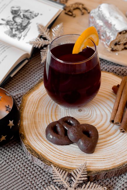 Glass of warm red beverage with orange slice, served with chocolate pretzels on a wooden tray, perfect for cozy winter setting.
