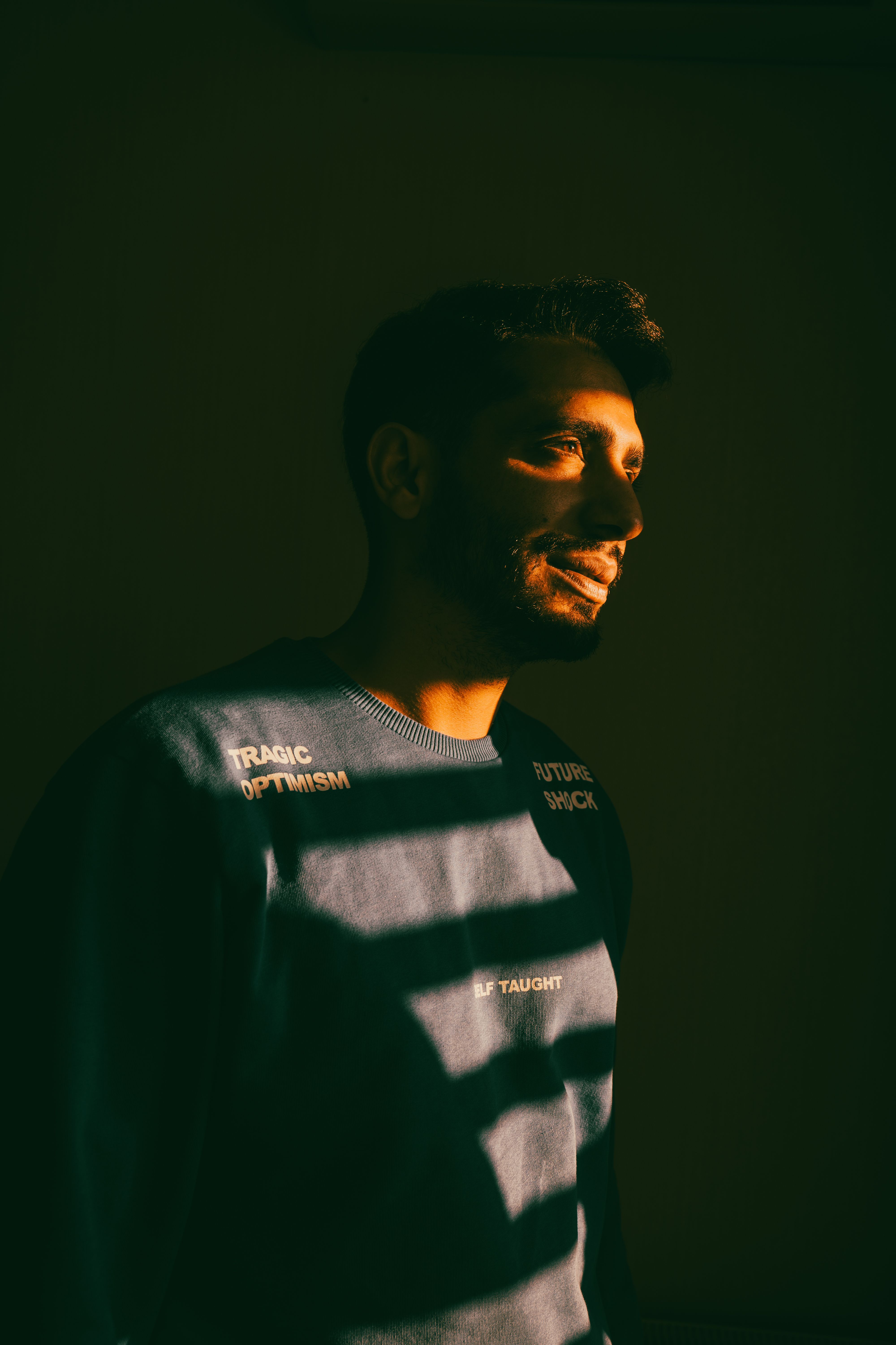 A man in a sweater with sunlight casting shadows through blinds, creating a moody portrait.