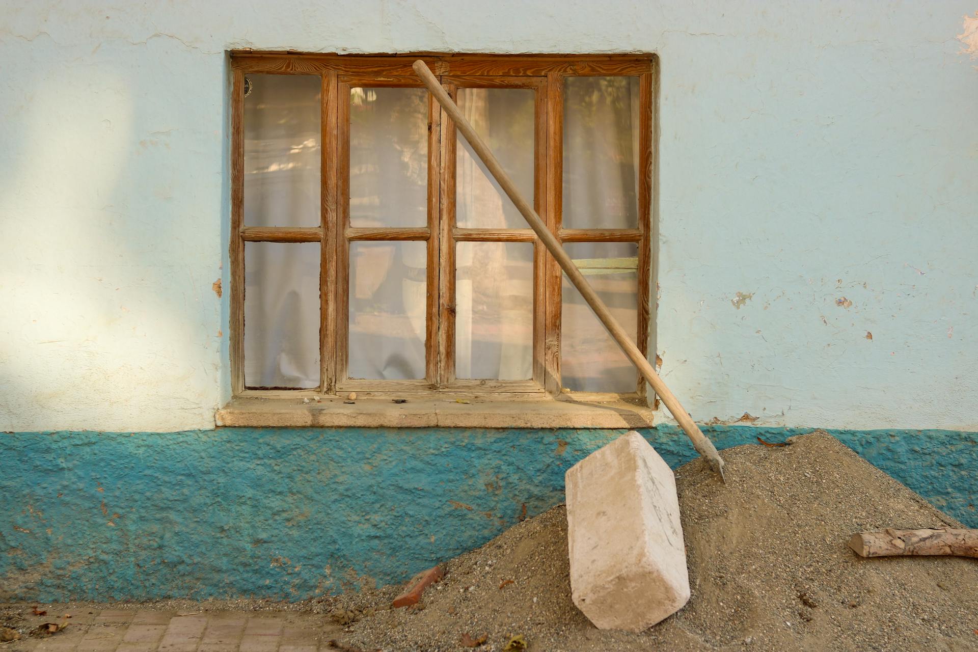 Old window on blue plaster wall with sand and gravel in sunny light. Construction materials lean against it.