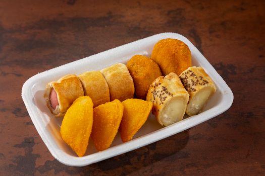 Tasty assortment of fried snacks including coxinha and sausage rolls on a foam plate.