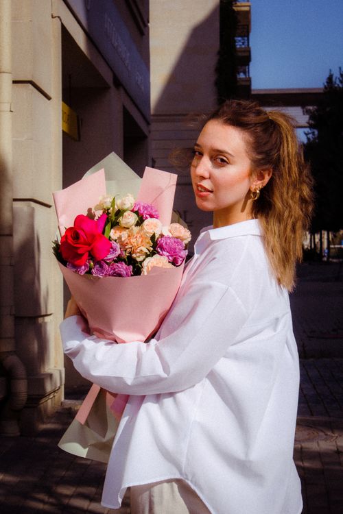 Free A young woman with long blonde hair holds a vibrant bouquet of flowers on a sunny day outdoors. Stock Photo