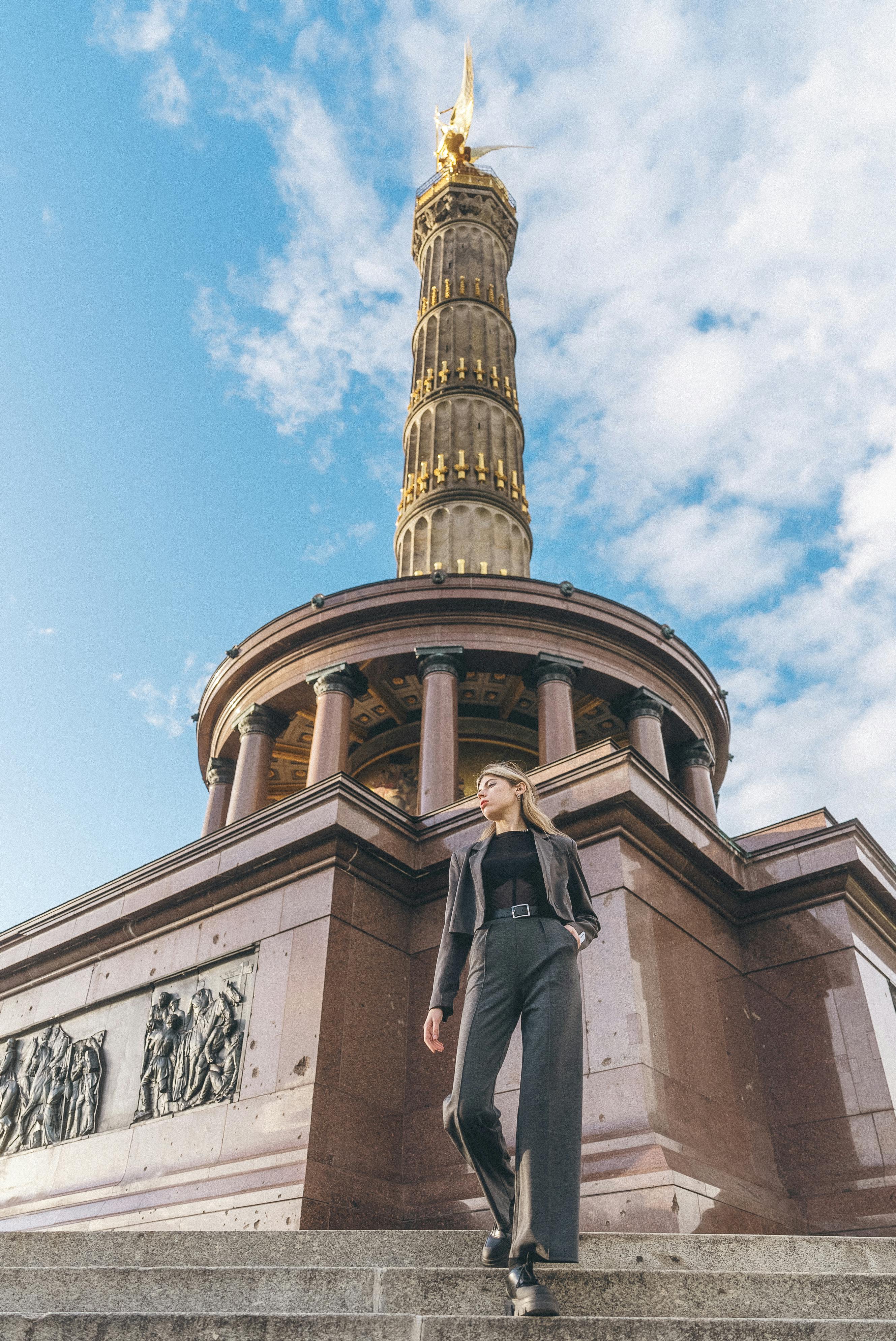 Free A confident woman poses at Berlin's iconic Victory Column, showcasing urban fashion style against a bright sky. Stock Photo