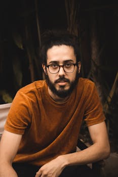 一位戴眼镜、留着胡须的时尚男子随意坐在户外，肖像如此