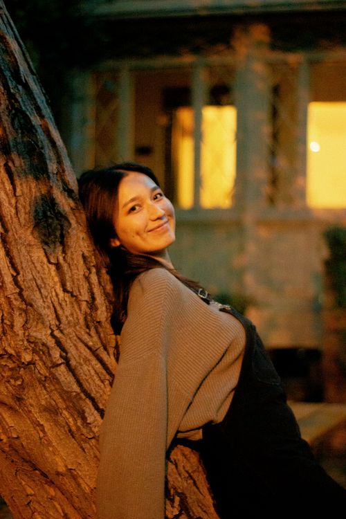 Free A cheerful woman leans against a tree outside at night, illuminated by warm light. Stock Photo