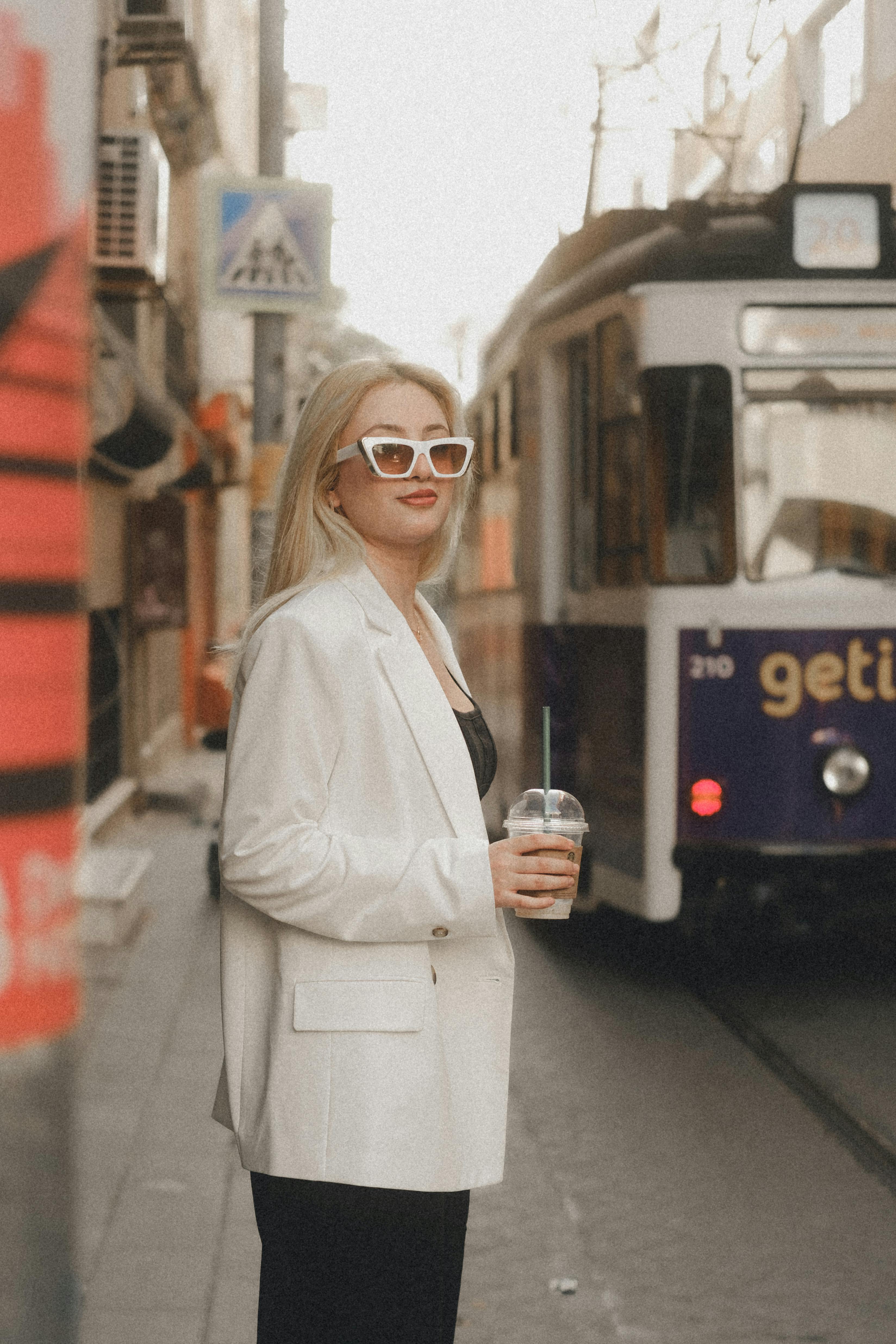 woman in white blazer with a coffee cup waiting on a sidewalk for the tram to pass