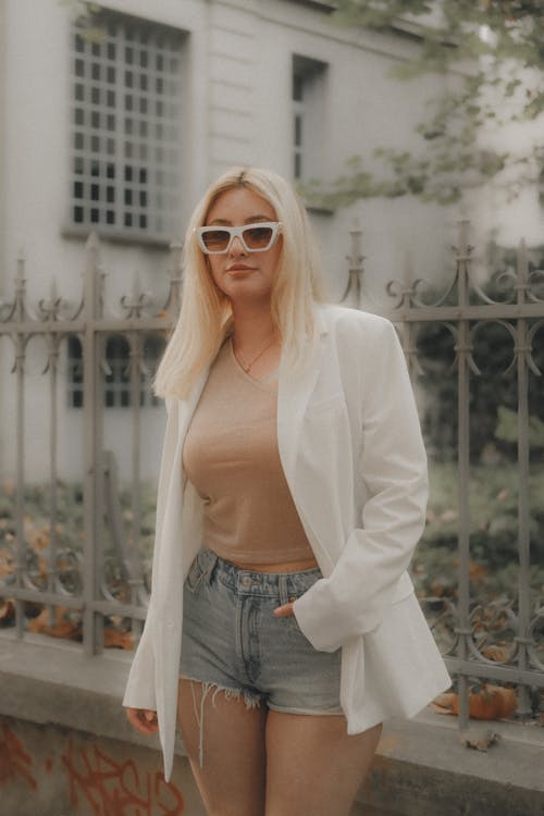 Model Wearing a White Blazer on Beige Blouse and Denim Cutoffs by the Fence