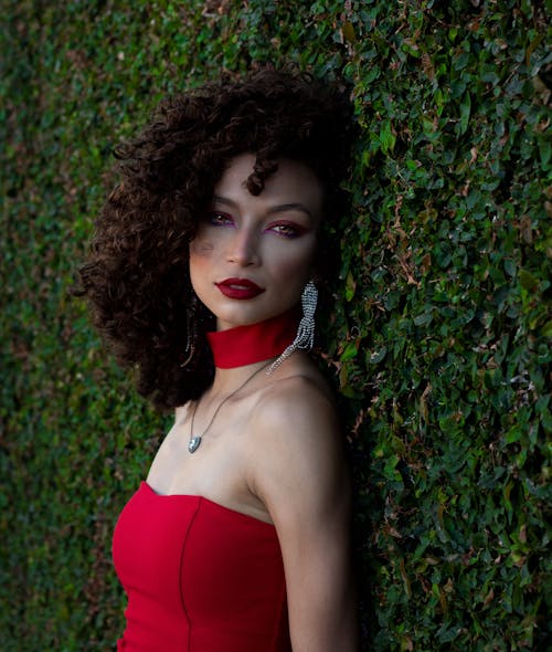Elegant Woman in a Red Dress and Red Lipstick Posing Outside next to a Shrub 