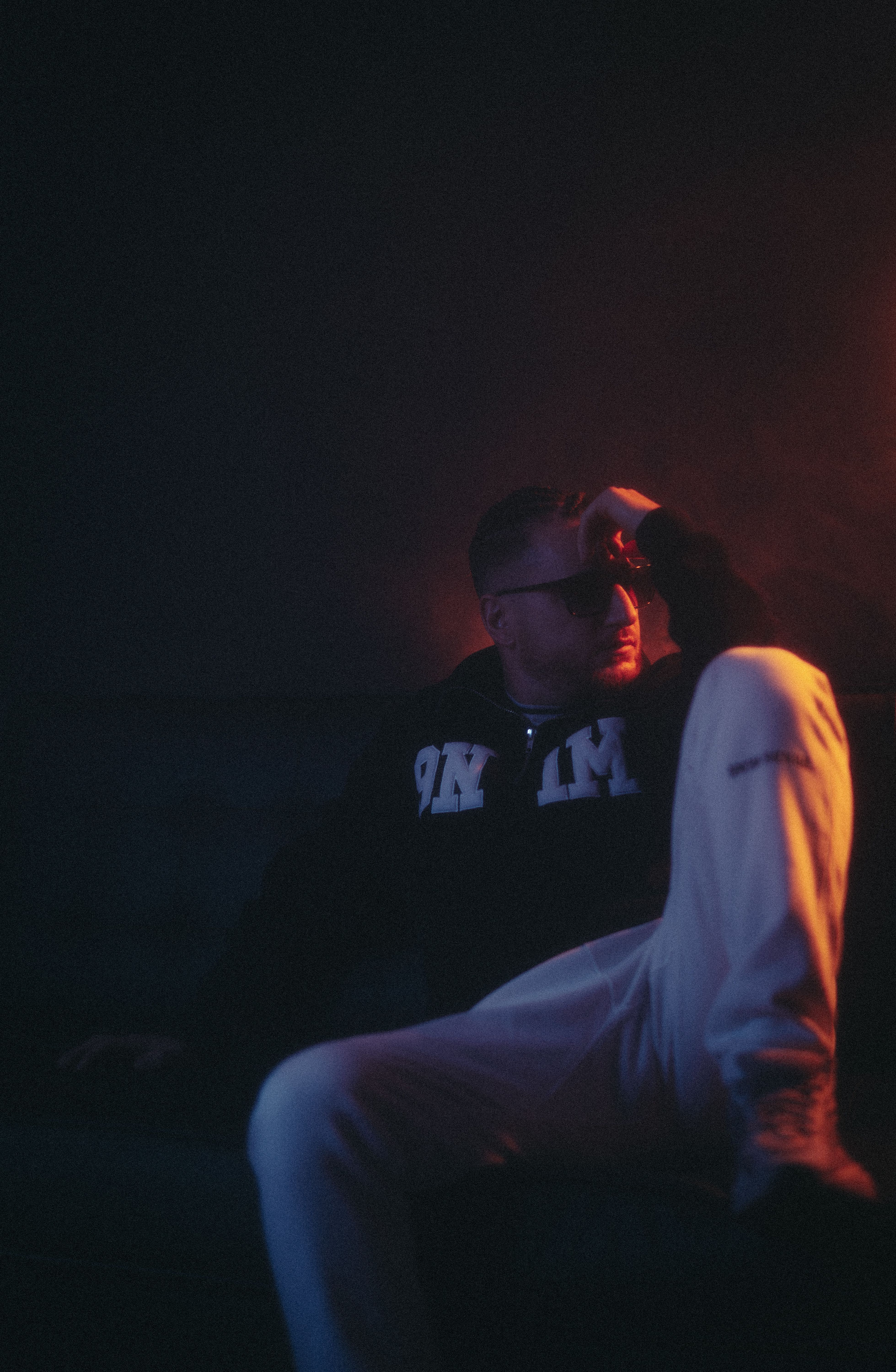 Fashion portrait of a man sitting in a dimly lit setting wearing eyeglasses.