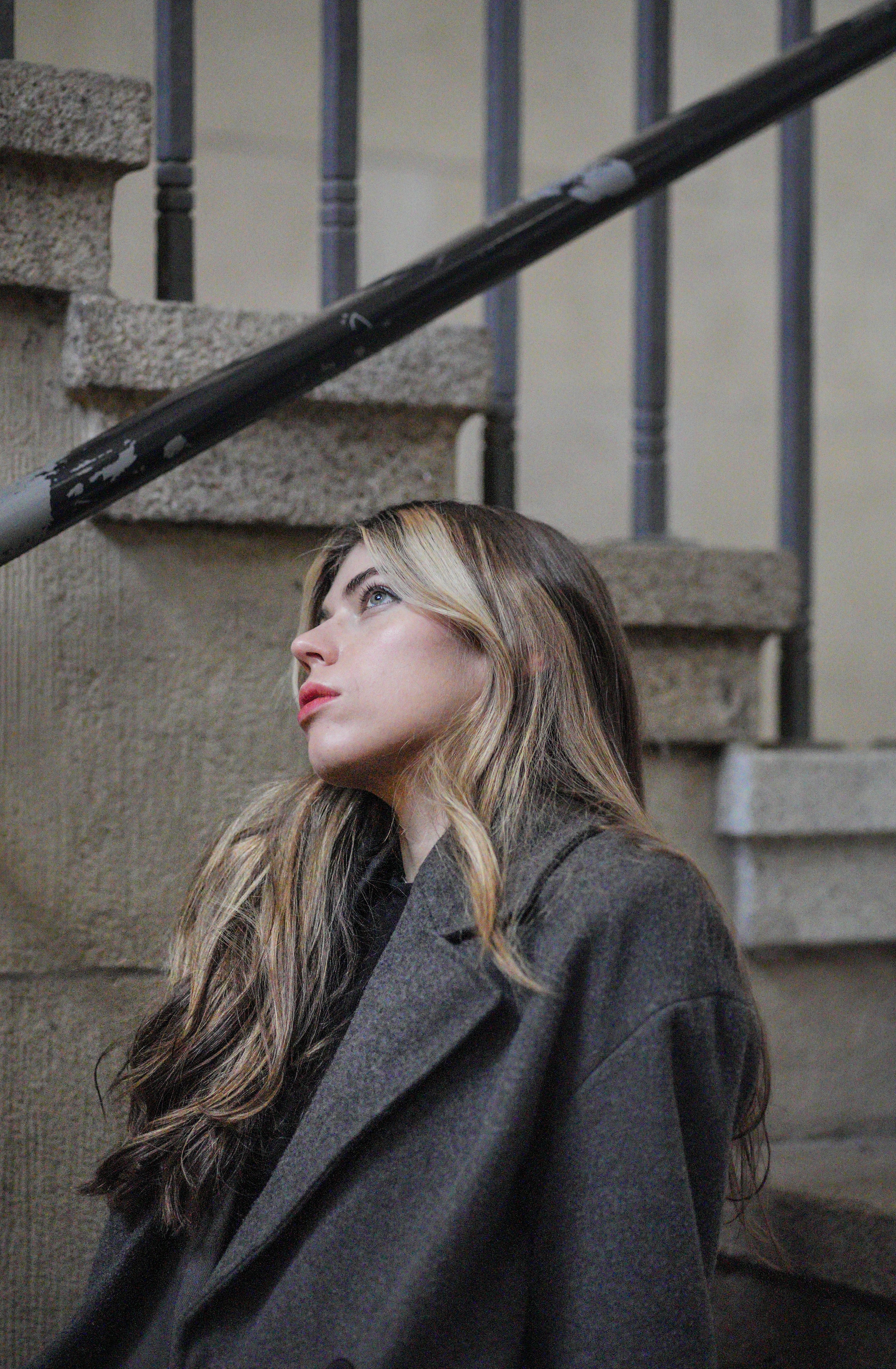 Young woman in fashion gray coat seated outdoors on urban stairs, looking contemplative.