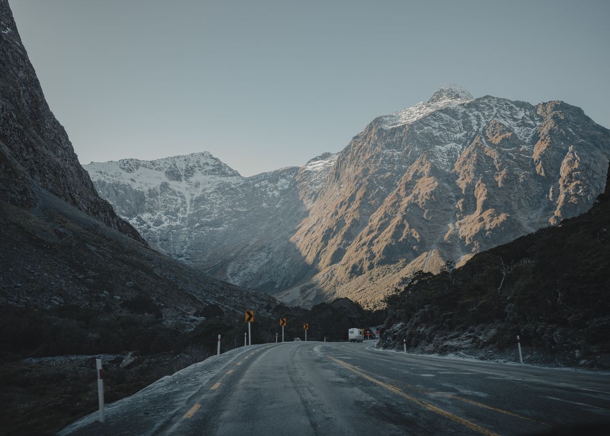 An empty alpine highway at dawn winding toward snow-dusted mountains, a single camper van visible in the distance