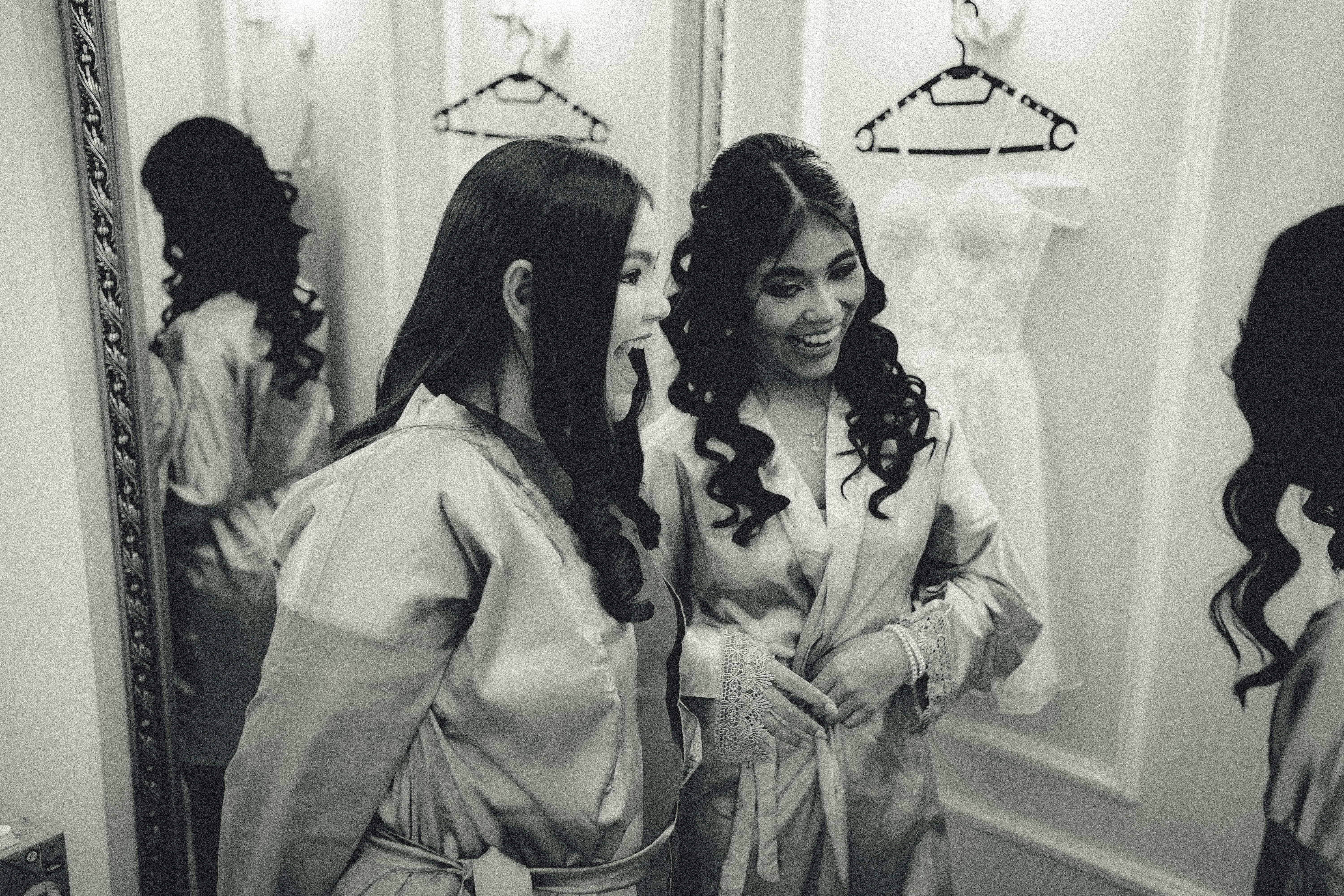 Two women in satin robes share a joyful moment in a dressing room before a ceremony.