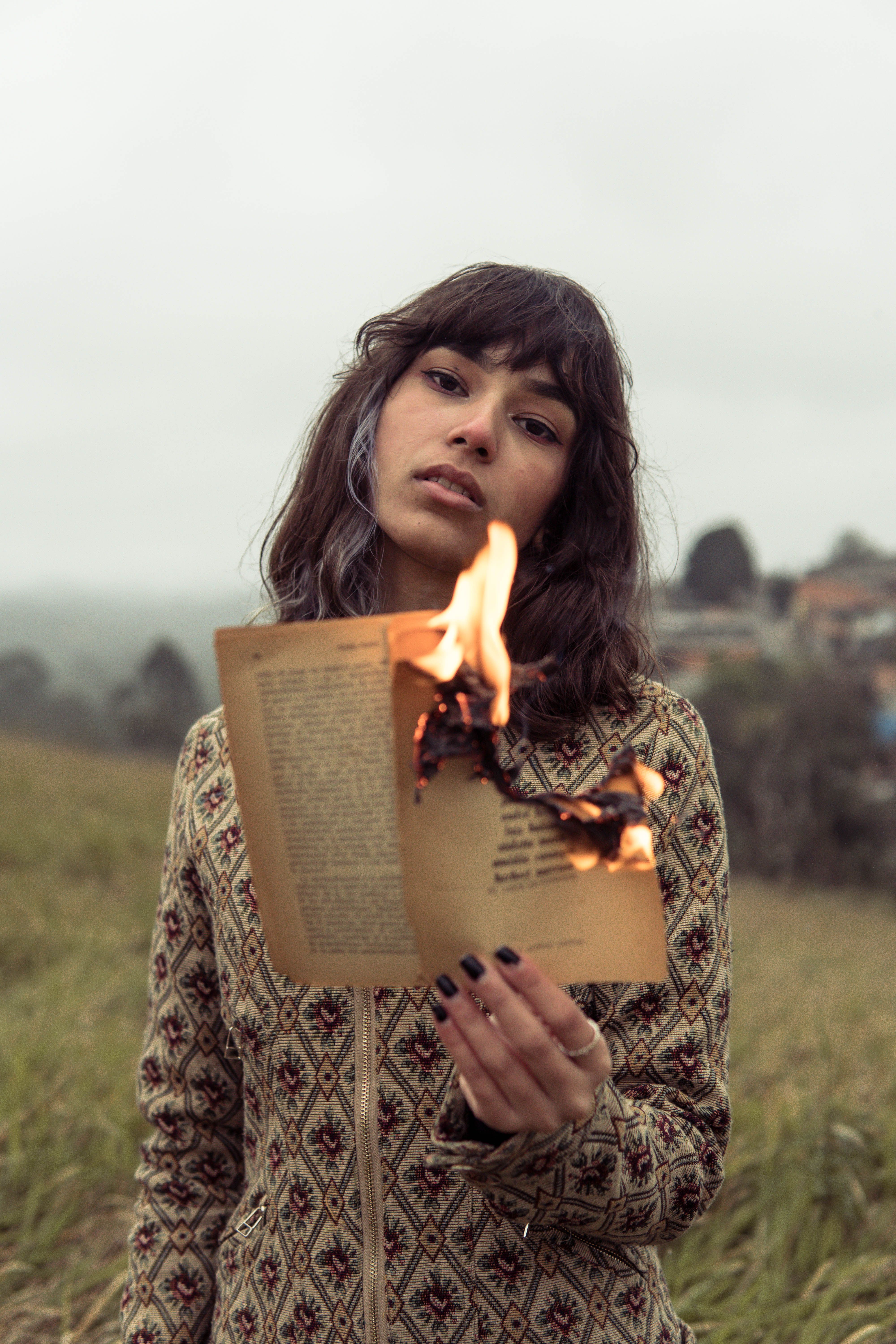 A woman holds a burning book in a misty field, blending fashion with dramatic storytelling.
