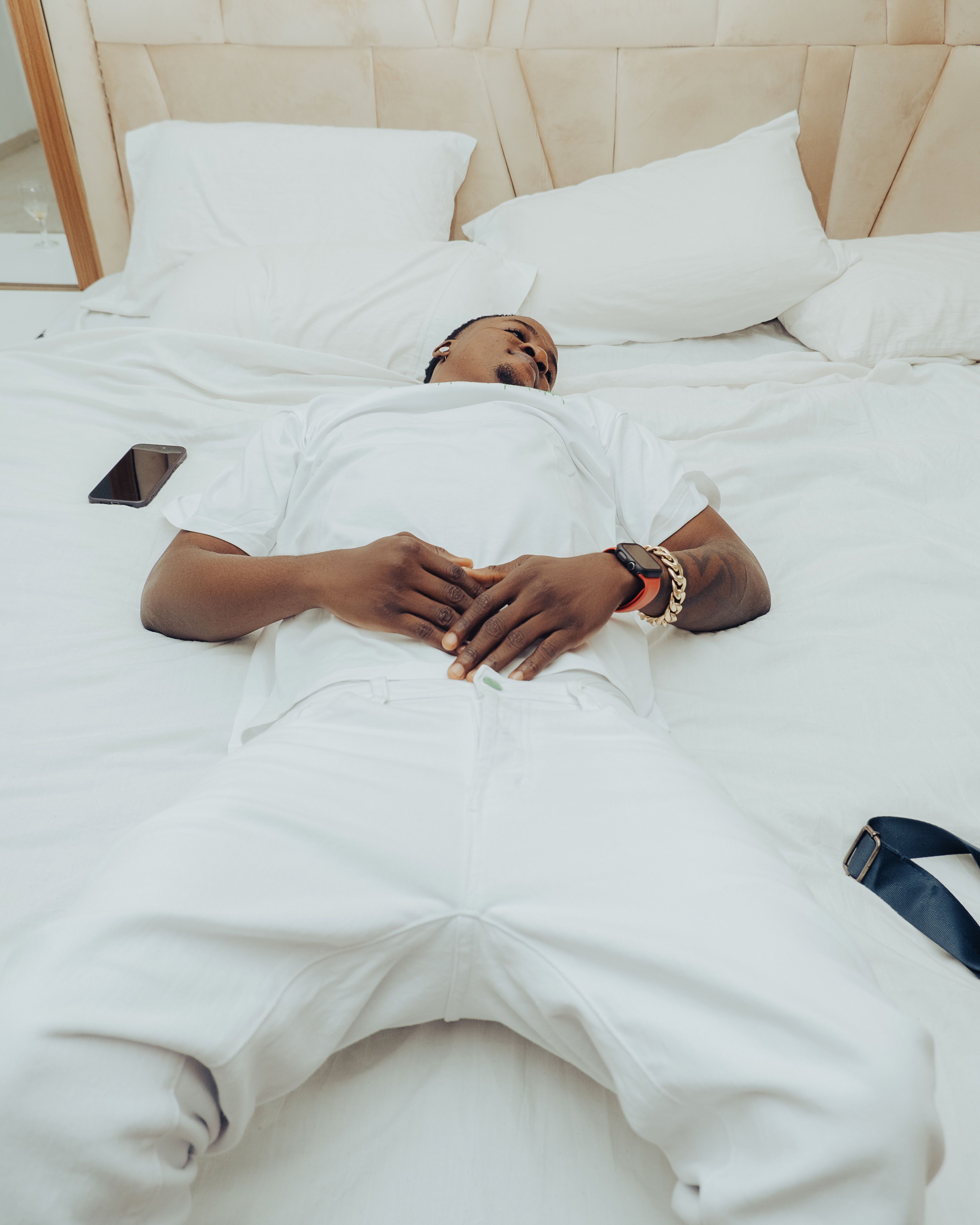 A young man lies on a white bed in a modern bedroom with relaxed posture and minimalistic decor.