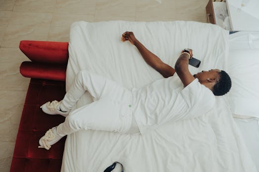 Young man in white outfit lying on bed in minimalist bedroom using smartphone from above angle.
