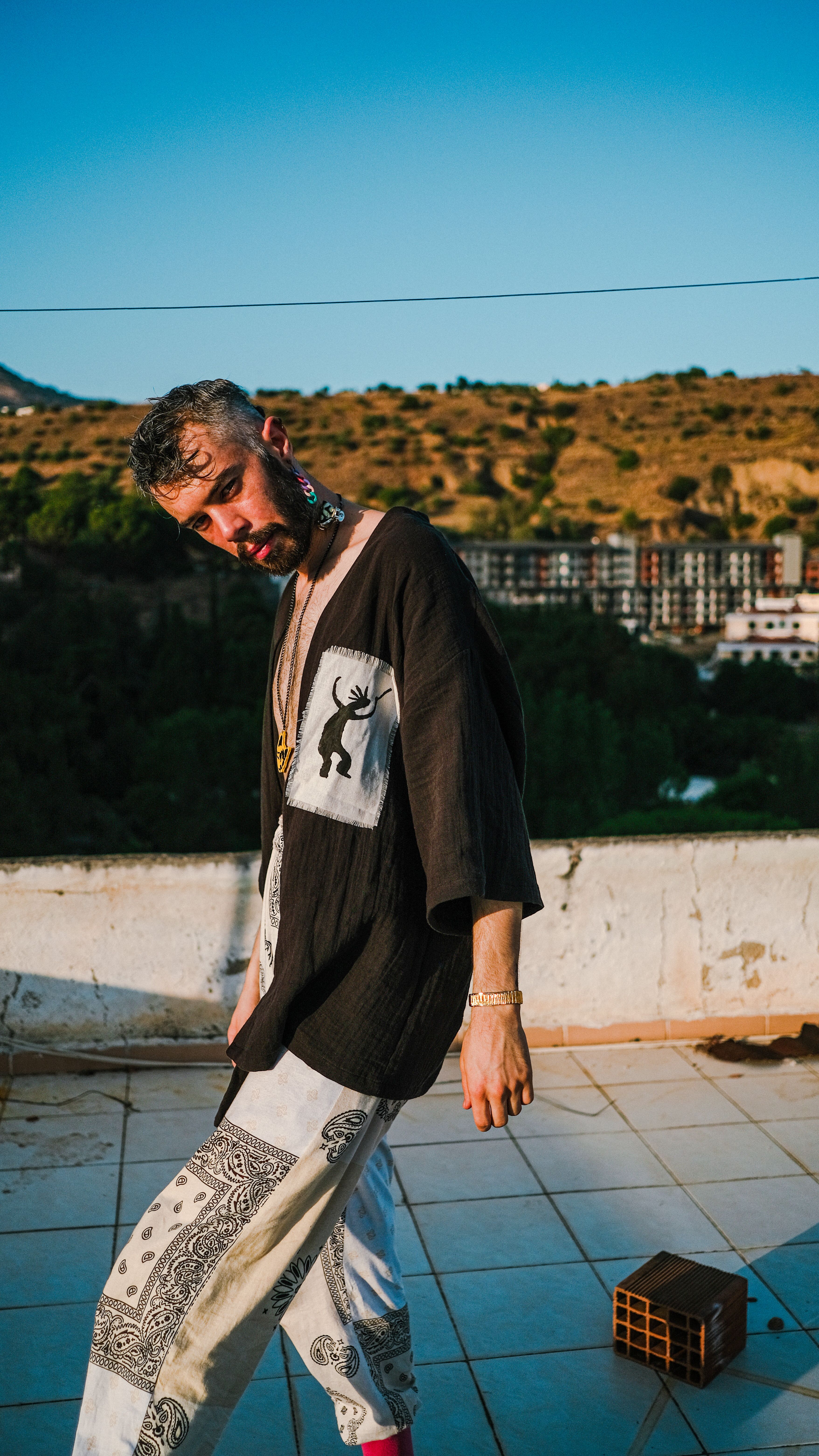 Fashionable young man with beard posing on a rooftop under vibrant blue sky.