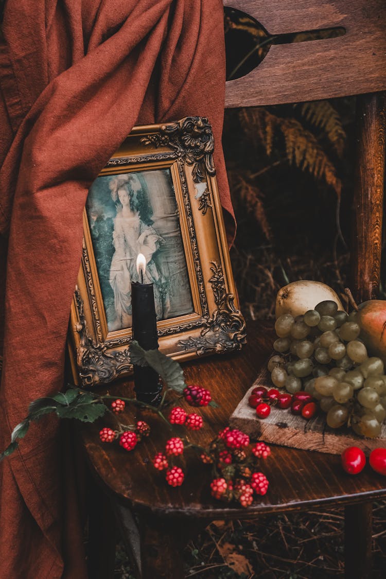 Old Fashioned Picture In A Decorative Frame Next To A Lit Candle And Fruits On A Chair