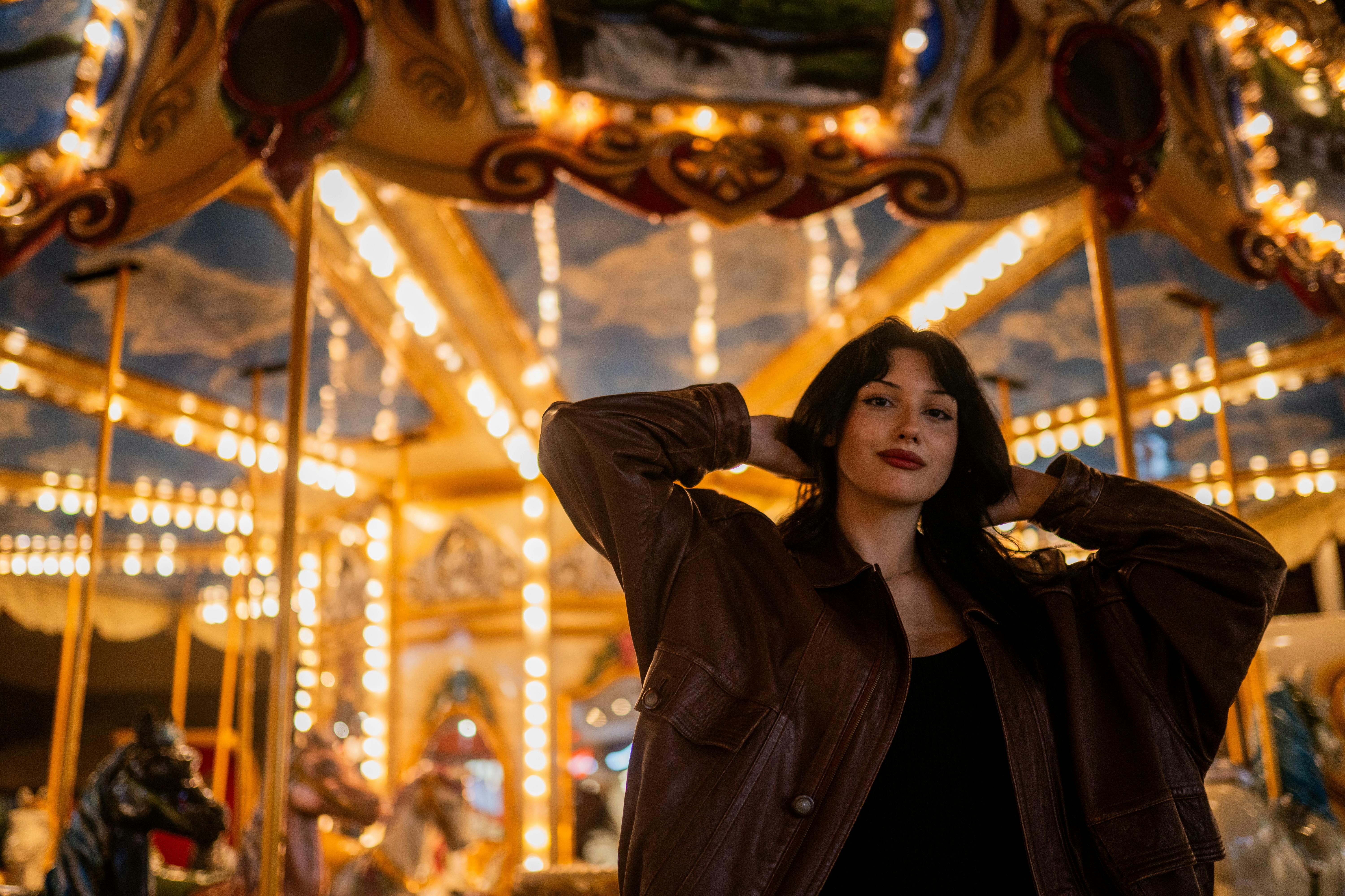 A young woman posing confidently at a beautifully illuminated carousel at night.