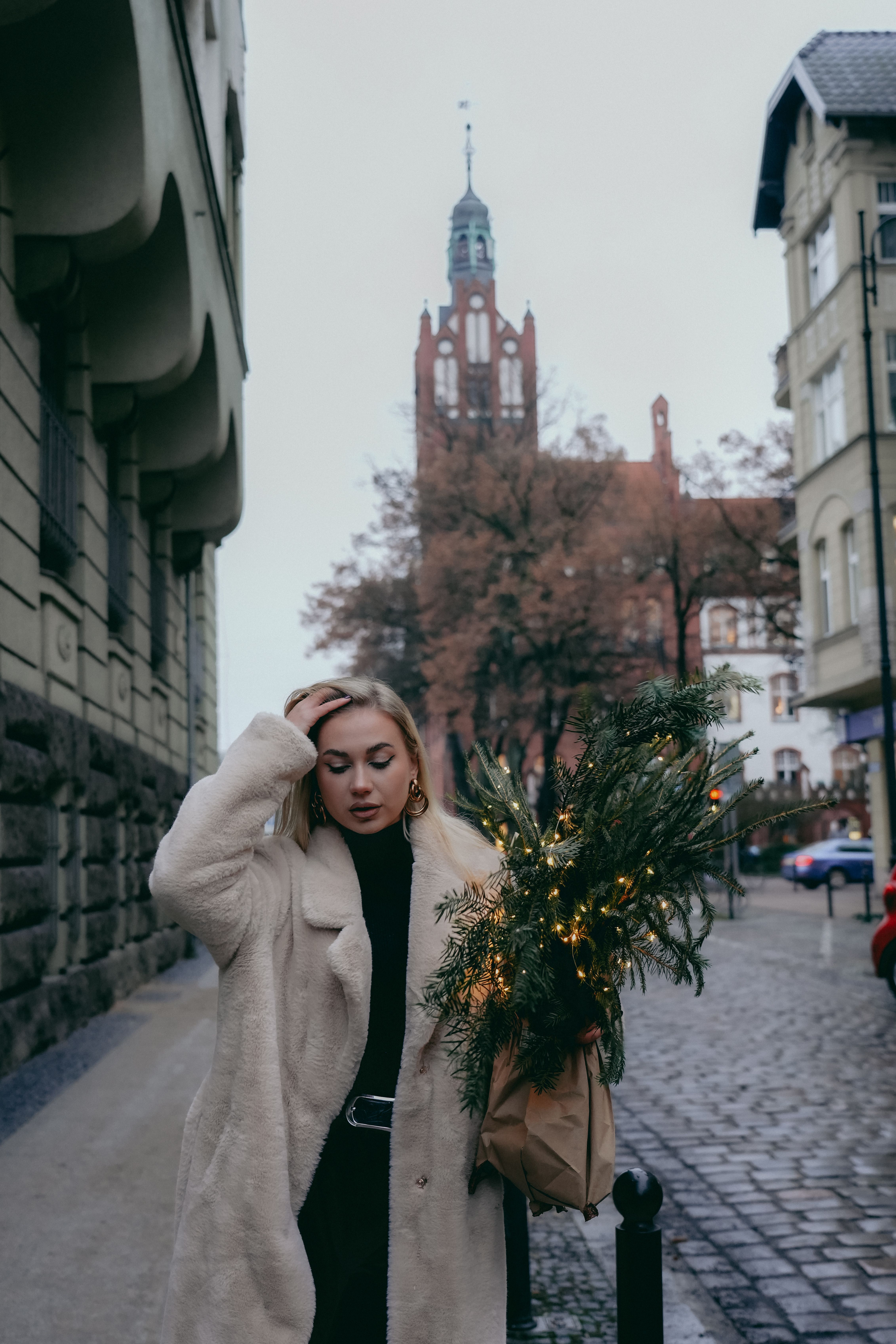 Woman in a fur coat carrying a bouquet walks through an old town street with historical architecture.