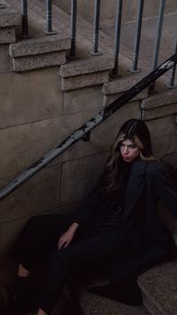 A fashionable woman in a black coat sitting on a stone staircase, exuding a dark and moody vibe.
