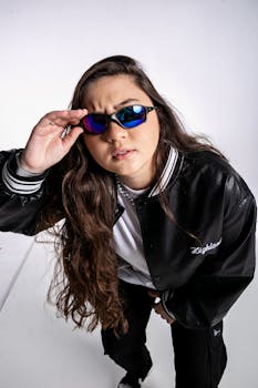 Fashionable young woman posing with sunglasses and leather jacket in studio.