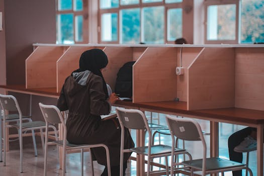 A female student wearing a hijab studies alone at a library desk, focused on learning.