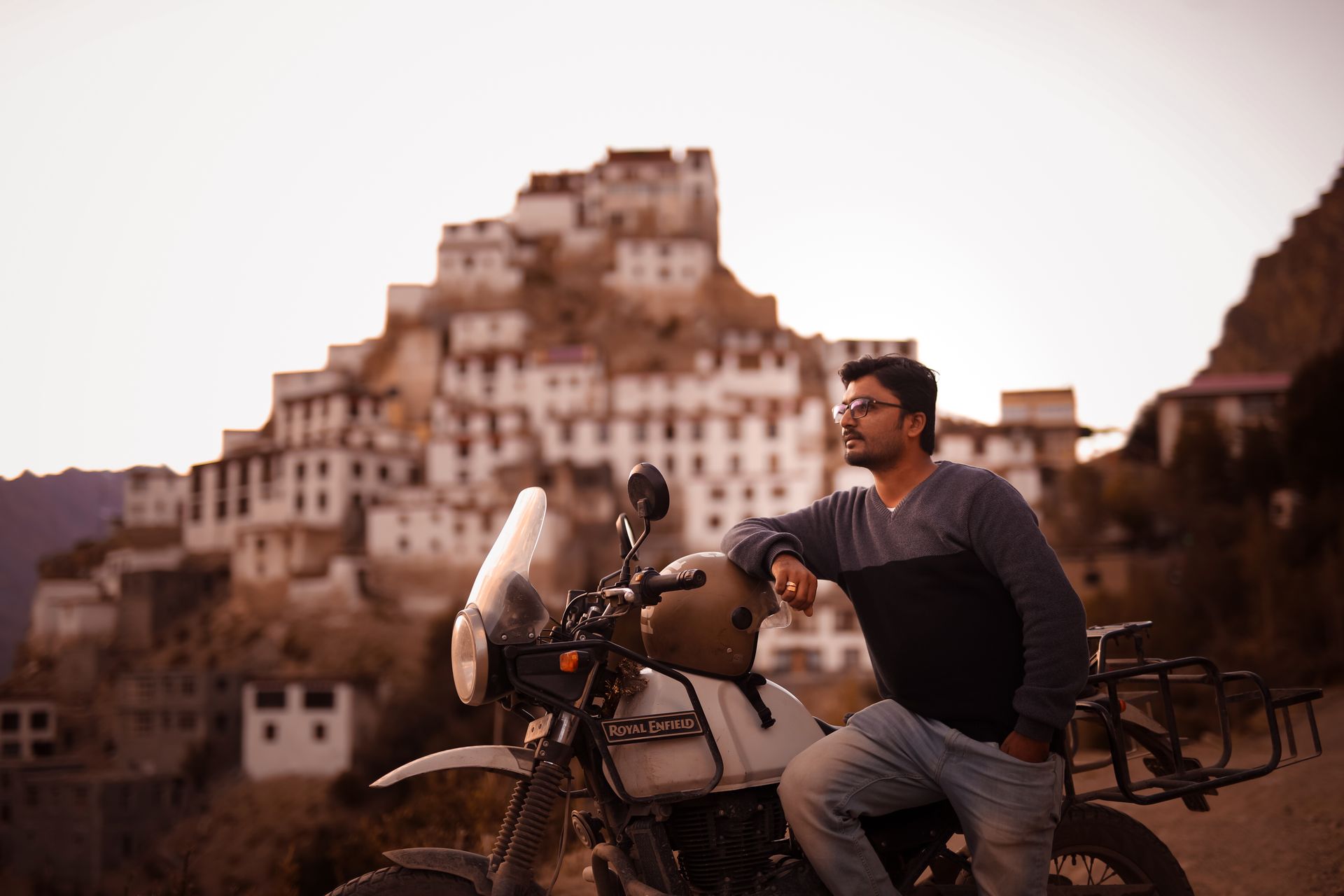 Man leaning on a Royal Enfield motorbike with a backdrop of Key Monastery in HP, India.