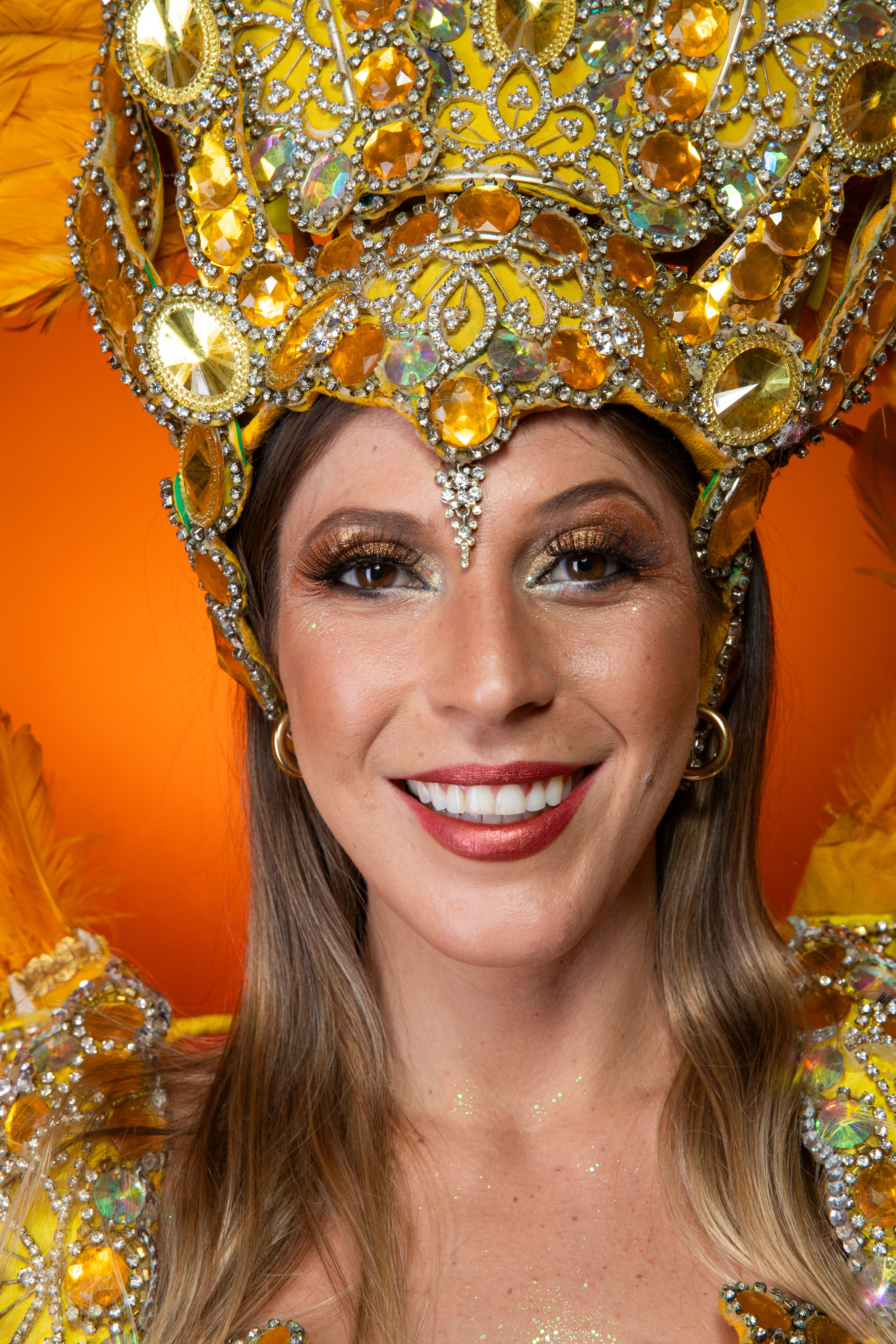 Colorful portrait of a smiling woman in a sequined carnival costume with an ornate crown on an orange background.