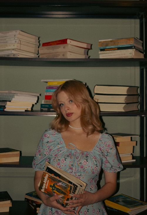 Free Stylish woman in a sundress holding books, standing in a library with shelves of stacked literature. Stock Photo