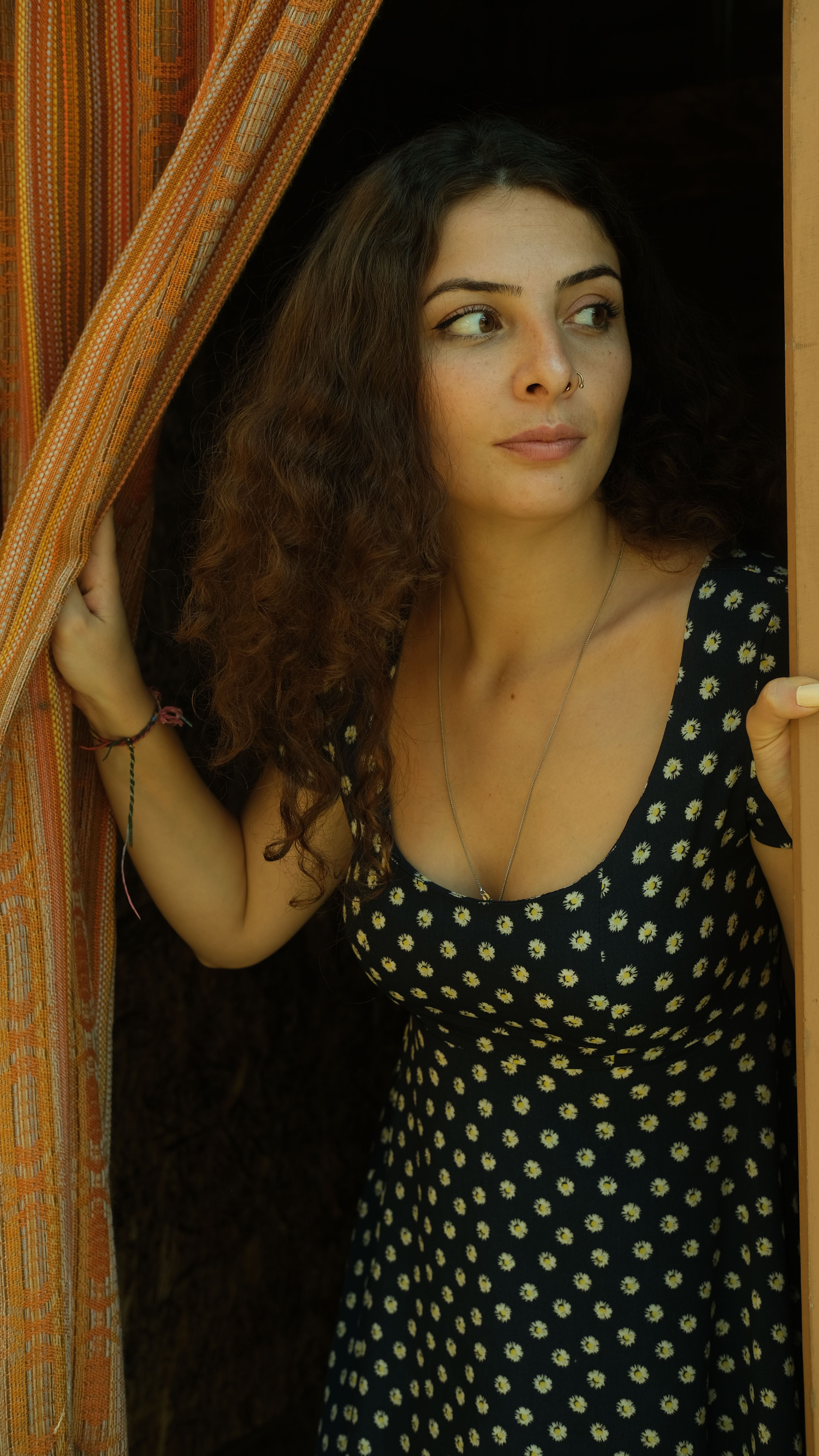 Portrait of a curly-haired woman peeking out from behind a curtain indoors.