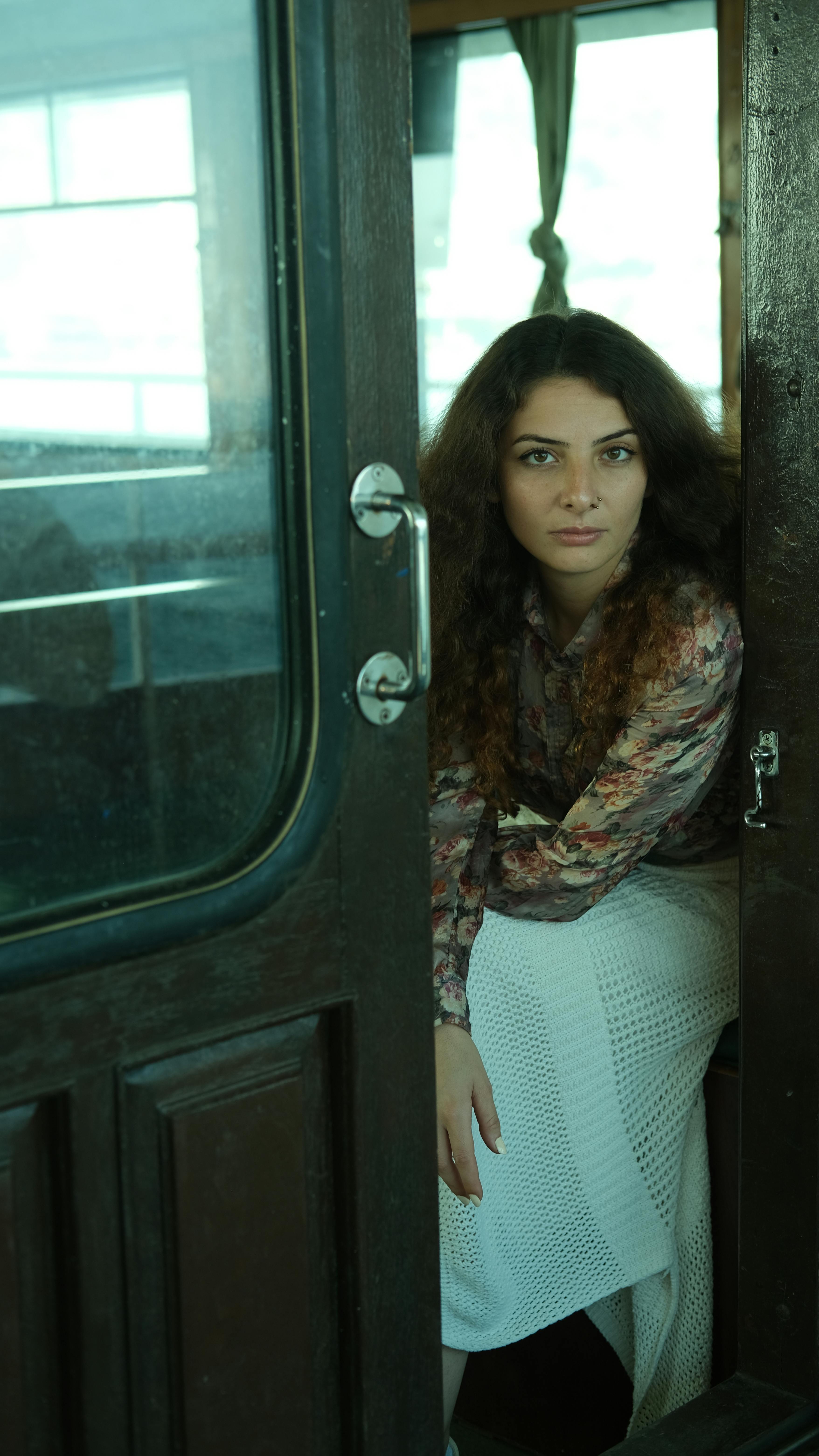 Young woman in floral blouse and white skirt sitting elegantly in a wooden doorway, creating a classic portrait.