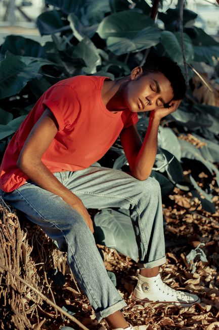 A young man in casual fashion rests outdoors among green leaves and sunlight.