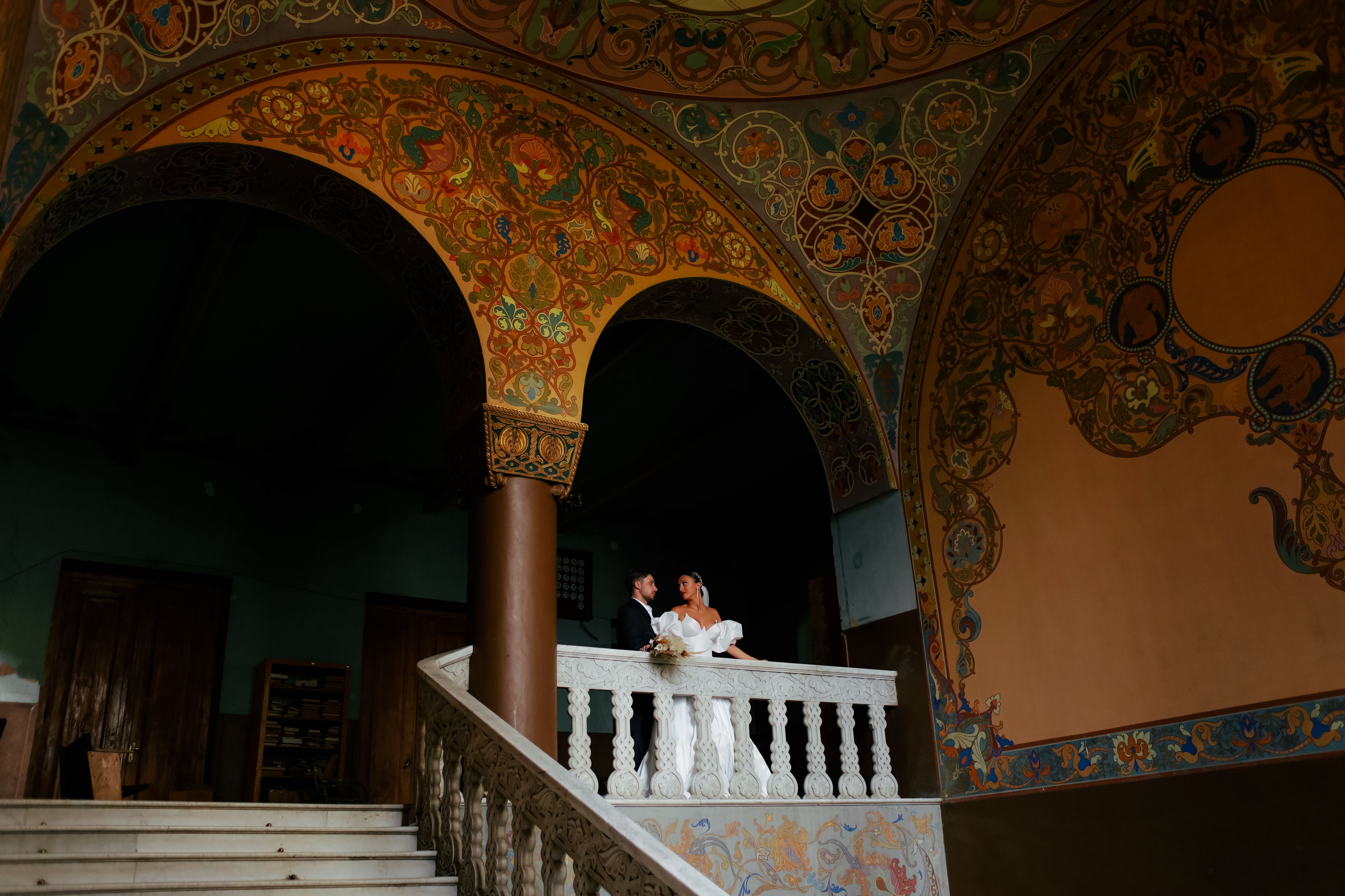 A young couple in wedding attire stands on a decorated historic staircase.