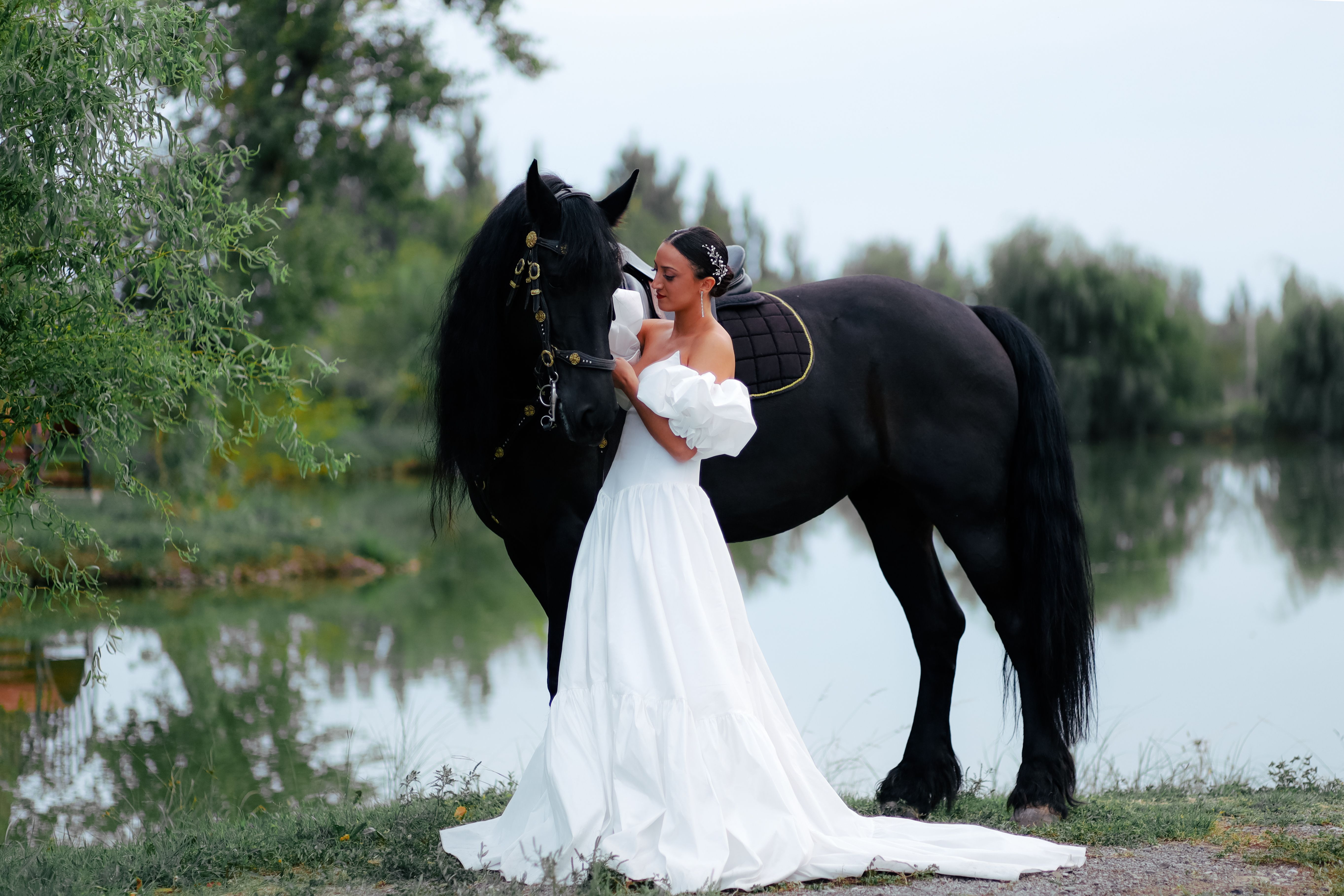 Elegant bride with black horse by a serene lake, capturing a beautiful outdoor wedding moment.