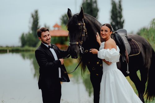 Free Bride and groom posing with a black horse by a serene pond. Stock Photo